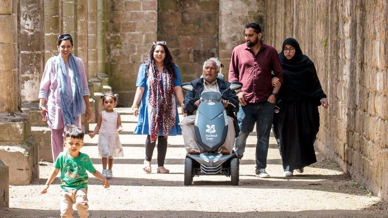 Large family including man on mobility scooter wandering beneath the columns of the nave in the abbey ruins with the sun shining through casting shadows.